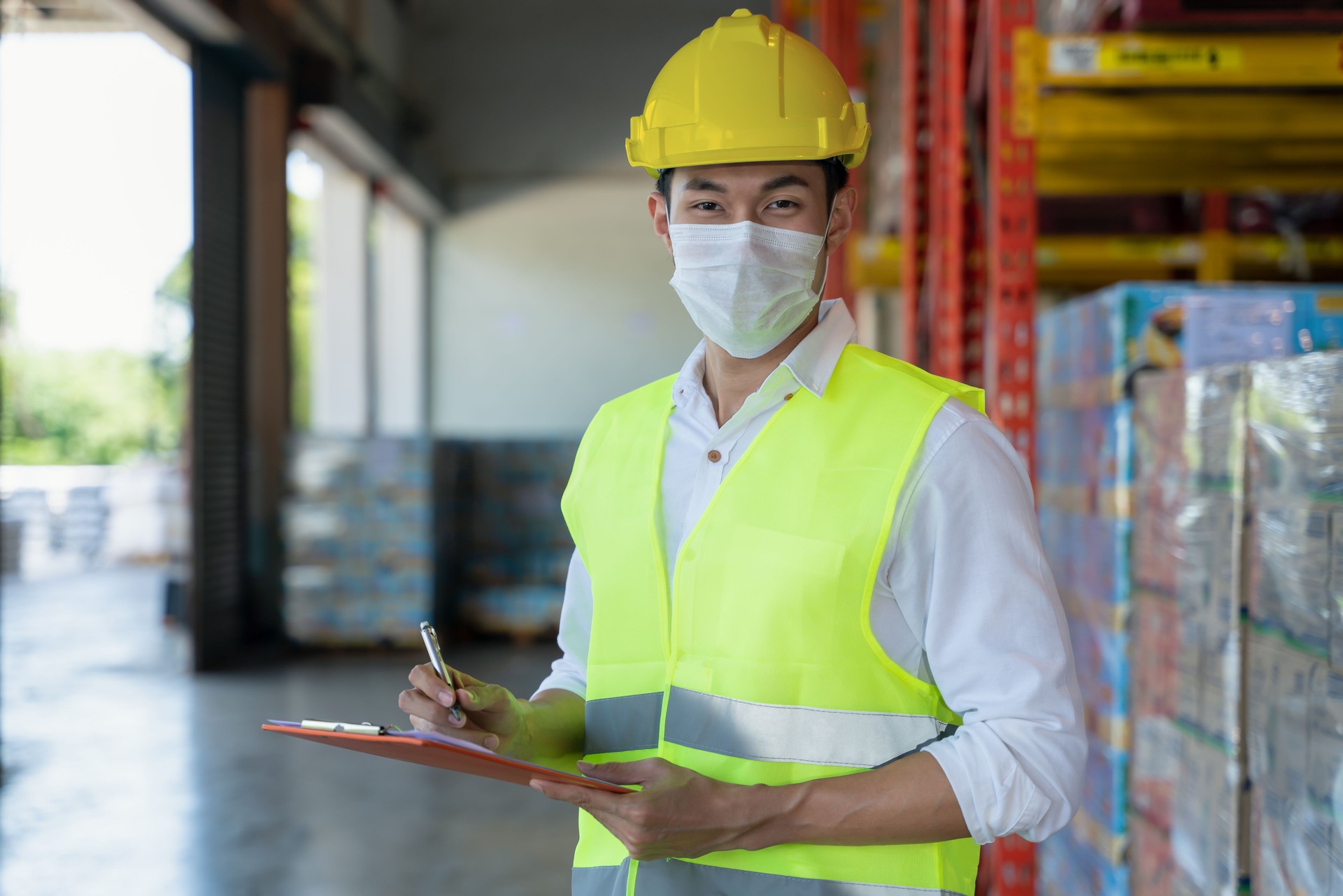 porttrait of asian warehosue woker with safety vest and helmet wearing medical mask to protect coronavirus covid-19 holding stock checking report at warehouse